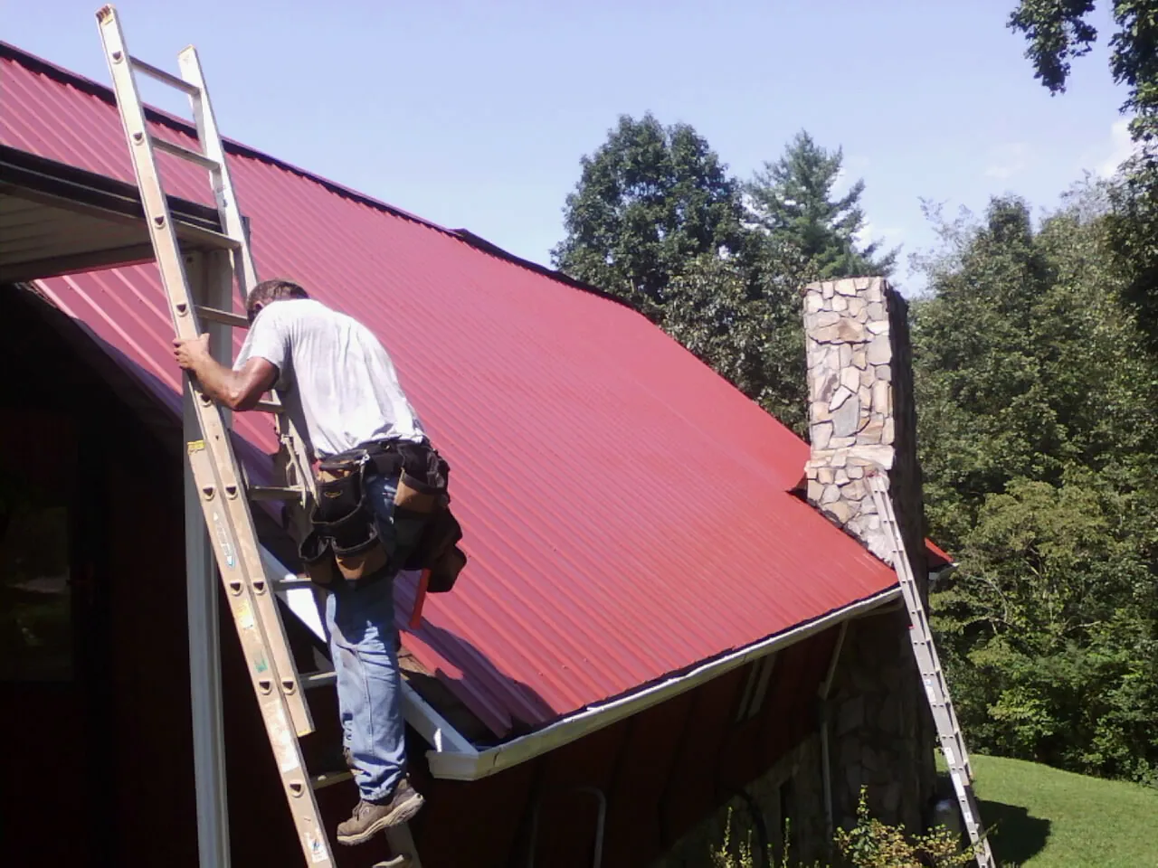 Roofer on a ladder beside a red metal roof and stone chimney — Soffit & Fascia Repair by Roofing Brothers in Thompson